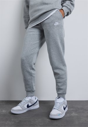 Personne portant un pantalon de jogging Nike gris et des baskets Nike blanches avec le logo noir, debout sur un sol gris devant un mur gris clair.