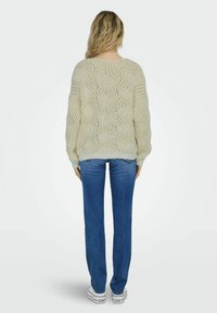 Cremig gestrickter Pullover mit Wellenmuster, lässiger Passform, Rundhalsausschnitt und gerippten Bündchen. Kombiniert mit blauen Jeans im geraden Schnitt und weißen Turnschuhen.