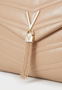 Borsa a tracolla beige in ecopelle trapuntata con accessori metallici in tonalità oro e un tassello a catena. Presenta un evidente dettaglio di chiusura a forma di V.
