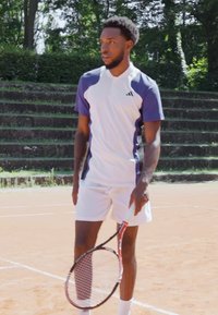 Tennis speler in een wit en blauw sportshirt met korte mouwen en witte shorts, die een tennisracket vasthoudt op een gravelbaan.