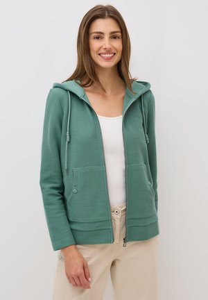 Green zip-up hoodie with a textured fabric, drawstring hood, two front pockets, and a casual fit, worn over a white shirt and beige pants.