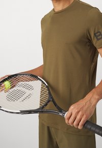 Olivově zelené sportovní tričko s krátkými rukávy, hladká textura. Osoba drží černý tenisový raket s žlutým míčkem, s přecházejícím vzorem.