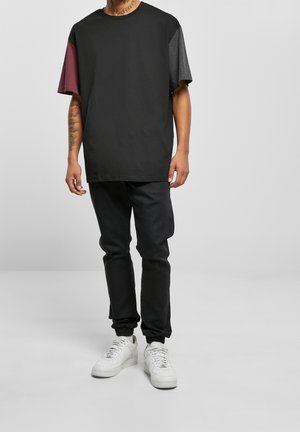 Persona con pantalones negros y una camiseta negra extragrande con mangas cortas granate y grises, de pie con zapatillas blancas sobre un fondo liso.