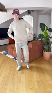 Felpa beige con tasca a marsupio, jeans azzurri, e sneakers argentate. Lo sfondo presenta un divano marrone con texture e una pianta in vaso.