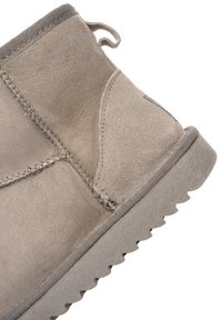 Primo piano di uno stivale invernale beige in pelle scamosciata con cuciture visibili, un cinturino in tessuto e una suola in gomma testurizzata.