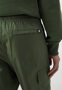 Olivgröna cargo-byxor med elastisk midja, med en bakficka och New Balance-logotagg. Tyget har en jämn textur.