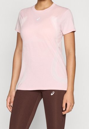 Vrouw draagt een lichtroze sportshirt met korte mouwen en donkerbruine legging met witte ASICS-logo's op borst en dij.