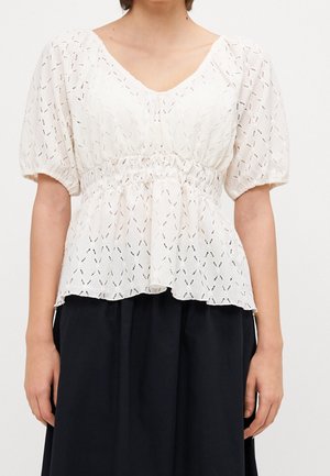 Nainen pukeutuneena valkoiseen, tekstuurilliseen lyhythihaiseen puseroon, jossa on poimutettu vyötärö, yhdistettynä mustaan hameeseen, yksinkertaisen taustan edessä.