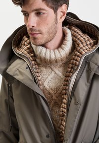 Giacca impermeabile verde oliva con cappuccio, aperta per rivelare un maglione beige a trecce e una fodera marrone a scacchi. Materiali testurizzati visibili.