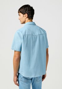 Camicetta in denim azzurro chiaro a maniche corte con colletto classico, dettaglio della spalla posteriore e vestibilità relaxed. Abbinata a jeans blu coordinati.