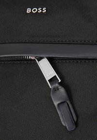 Schwarze Stofftasche mit einem silbernen Reißverschluss-Zipper, der einen Lederanhänger hat. Das „BOSS“-Logo ist über dem Reißverschluss in den Stoff geprägt.