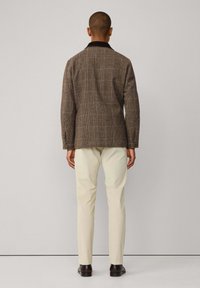 Bruine geruite jas met een zwarte kraag, gecombineerd met off-white broek en donkere schoenen. De stof lijkt textuur te hebben en is op maat gemaakt.