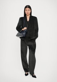 Femme debout portant un costume noir texturé avec un pantalon évasé, des talons noirs et portant un sac à épaule bleu marine.
