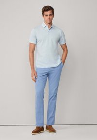 Man som står med ena handen i fickan, iförd en ljusblå pikétröja, blå byxor och bruna mockaskor, framför en enfärgad bakgrund.