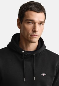 Sort pullover-hoodie lavet af fleece-stof. Har en hætte med snor og et lille broderet logo ved brystet med sølvaccent.