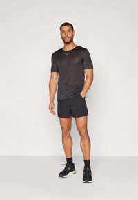 Svart trenings-T-skjorte med rund hals og korte ermer, kombinert med svarte shorts. Har et logo på brystet og bæres med svarte joggesko.