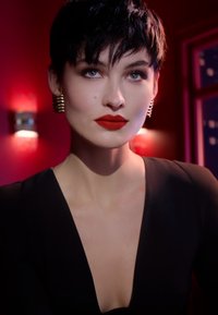 Robe noire à décolleté en V profond, cheveux courts et foncés, lèvres rouges éclatantes, boucles d'oreilles en spirale métalliques, peau douce, illuminée par un éclairage de fond rouge.