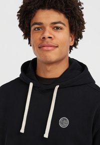 Haut à capuche noir avec un cordon de serrage, accents blancs et un patch logo circulaire sur la poitrine. Présente une coupe décontractée et une texture de tissu douce.