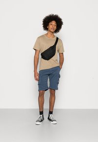 Zwarte heuptas gedragen over de borst, beige shirt met korte mouwen, blauwe cargoshorts en zwart-witte hoge sneakers. Eenvoudig ontwerp.