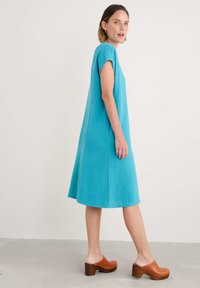 Turquoise katoenen jurk met korte mouwen, A-lijn vorm, textielstof, tot halverwege de kuit, gecombineerd met bruine houten klompen.