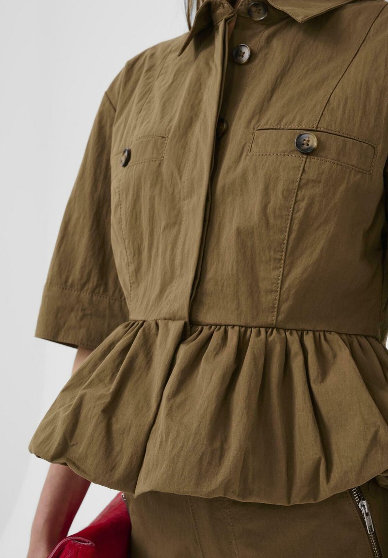 Bruine jasje met knopen, twee borstzakken en een geplooide peplum taille, gedragen met bijpassende broek en een rode clutch in de hand.