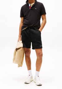 Svart polo-shirt med krage och mönstrad innersida; svarta sportshorts; beige sweatshirt som hänger över armen; vita sneakers.