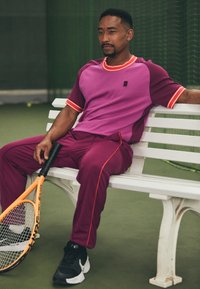 Completo sportivo composto da una maglietta a maniche corte magenta e viola con rifiniture arancioni, abbinata a pantaloni viola con dettagli rossi. Sneakers nere e una racchetta da tennis.