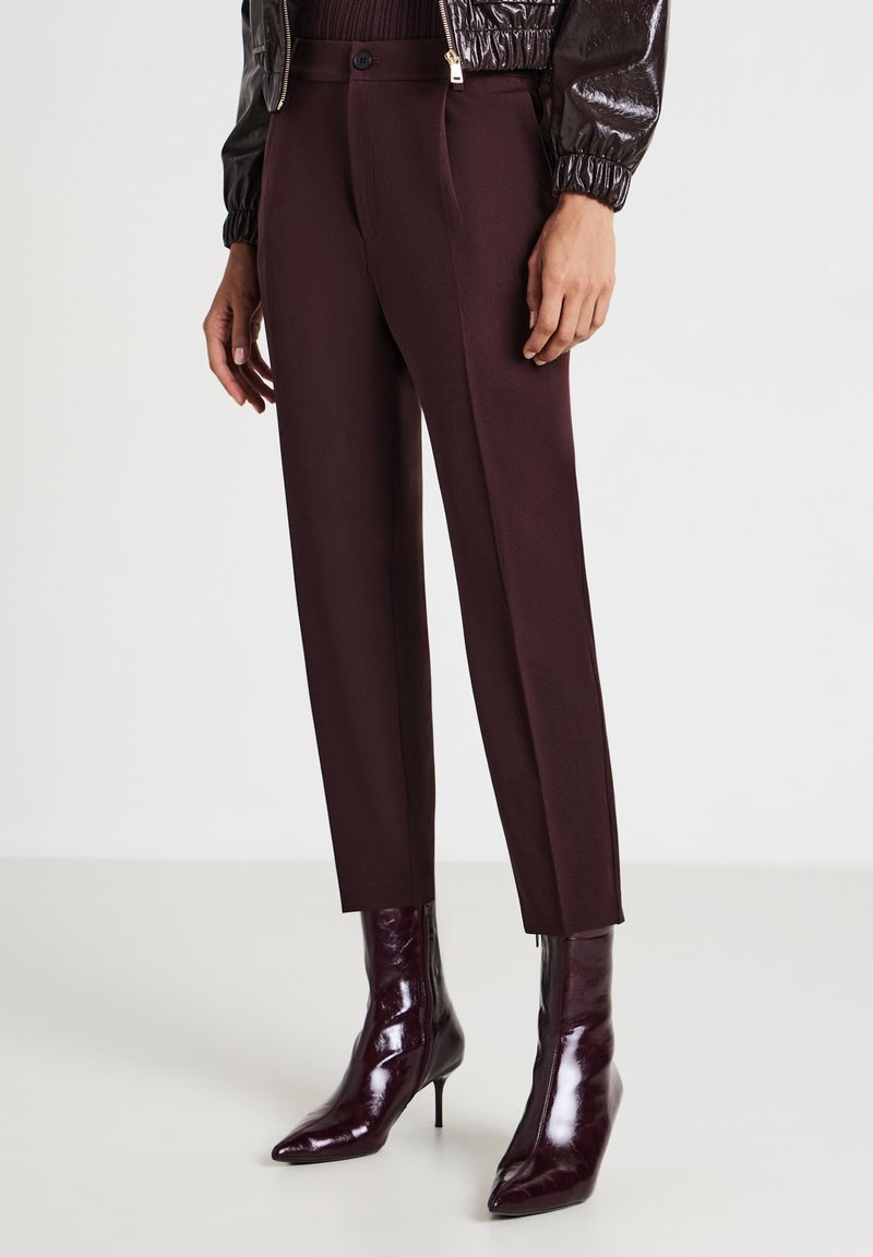 Femme portant un pantalon taillé taille haute bordeaux foncé et des bottes à talons aiguilles brillantes assorties, se tenant sur un sol blanc.