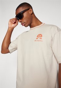 Camisa de algodón beige de manga corta con logo naranja y diseño de engranaje. Presenta un cuello redondo y dobladillo de contraste. Modelo lleva gafas de sol negras.