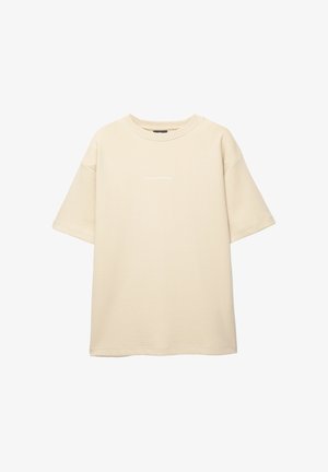 Beige ylisuurikokoinen lyhytnokkatoppi, joka on valmistettu pehmeästä kankaasta. Sisältää pyöreän kaula-aukon ja pienen tekstidetaljin etuosassa.