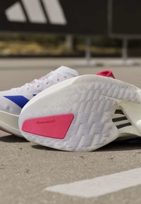 Λευκά αθλητικά παπούτσια Adidas για τρέξιμο με μπλε και κόκκινες πινελιές, που δείχνουν την σόλα με ροζ λογότυπο Continental σε υπαίθριο πεζοδρόμιο.