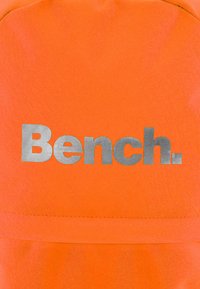 A narancssárga szövet sima textúrával rendelkezik, amelyen egy feltűnő ezüst "Bench." logó található. A dizájn egyszerű és modern, a márkázásra összpontosít.