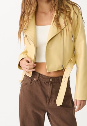 Femme portant une veste en cuir jaune pâle courte avec des détails zippés, sur un crop top blanc et un pantalon marron taille haute.