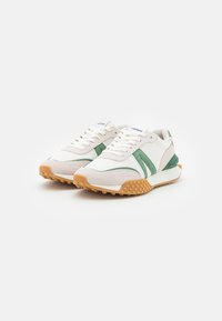 Des chaussures de sport blanches présentent des accents verts, un dessus en maille texturée et une semelle en caoutchouc gomme avec un design à clous pour une adhérence optimale.