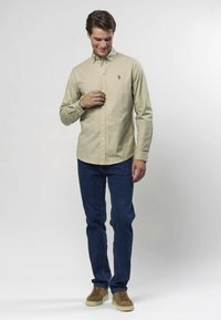 Hellbeiges Hemd mit Knopfleiste, Kragen und kleiner Stickerei auf der Brust, kombiniert mit dunkelblauen Jeans und hellbraunen Schuhen.