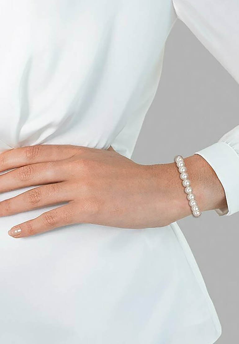Pulsera de perlas con perlas blancas redondas ensartadas en una banda elástica, llevada en una mano que descansa en la cintura de una blusa blanca.