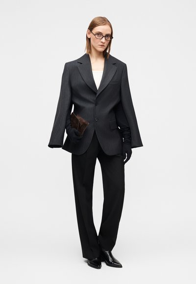 Veste de costume noire à rayures fines surdimensionnée avec manches larges, fermé par des boutons et grande poche. Portée avec un pantalon noir sur mesure et des bottes pointues.