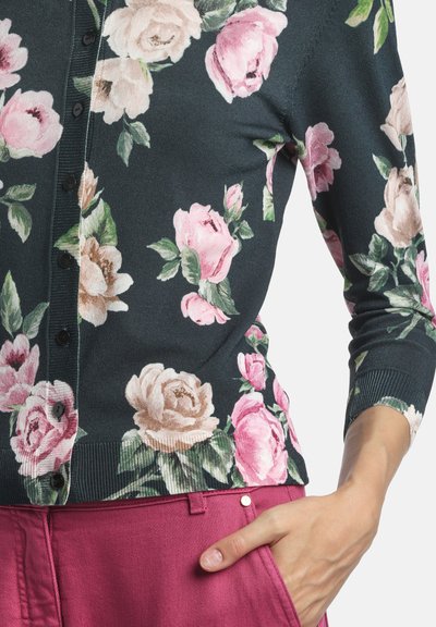 Cardigan à fleurs vert foncé orné de grandes roses roses et crème. Tissu en maille douce avec poignets côtelés et boutons sur le devant. Pantalon rose visible.