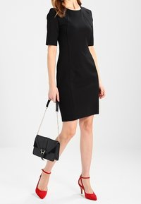 Musta muotoileva mekko lyhyillä hihoilla ja sauman yksityiskohdilla, yhdistettynä punaisiin remmiin korkeisiin kenkään ja mustaan käsilaukkuun ketjuhihnalla.