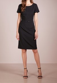 Robe noire à manches courtes avec une silhouette ajustée, jupe longueur genou, plis subtils à la taille et un détail plat au centre.