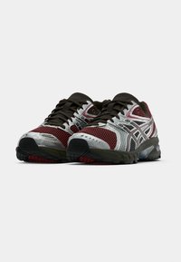 Piros és ezüst sportcipők hálós, szintetikus anyagból, fekete fűzőkkel és gumitalppal. Díszítő mintákkal és szellőző nyílásokkal rendelkeznek.