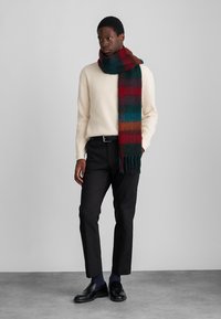 Écharpe en laine à rayures rouges, vertes et bleues avec des détails à franges, portée avec un pull texturé crème et un pantalon noir. Des chaussures lisses complètent la tenue.