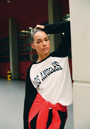 Femme aux cheveux tirés en arrière, portant un T-shirt Los Angeles et une veste rouge nouée autour de la taille, s’appuie contre un pilier à l’intérieur.