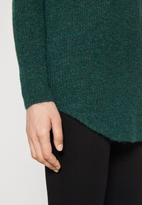Zielony sweter z dzianiny o textured wykończeniu, z zaokrąglonym dołem i długimi rękawami, w zestawie z czarnymi dopasowanymi spodniami.