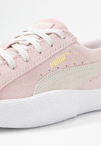 Vaaleanpunaiset nahkakengät, joissa on valkoiset nauhat, vaalean beige sisustus ja kuvioitu valkoinen kumipohja. Sivussa on kultainen logo.