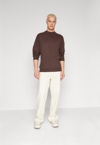 Suéter de punto marrón con cuello alto y puños acanalados, combinado con pantalones blancos de corte holgado y zapatillas de colores claros.