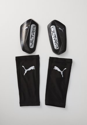 Mustat jalkapallosuojat "ULTRA Flex" -merkinnällä ja metallikoristeilla, yhdistettynä mustiin kangassukkiin, joissa on valkoinen Puma-logo.