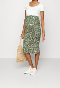 Maternity skirt featuring a green floral pattern, stretchy fabric, knee-length design, paired with a white fitted shirt and woven bag.