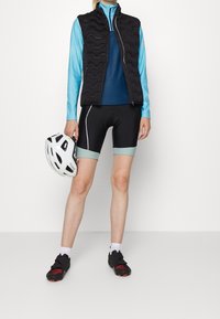 Person som bär svarta cykelshorts, blå långärmad topp, svart väst och svarta skor, håller en vit cykelhjälm vid sin sida.