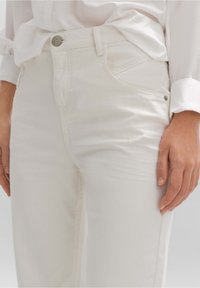 Jeans de mezclilla blanca con diseño ajustado, ligero desgaste cerca del muslo y botones metálicos en la cintura. Textura suave.